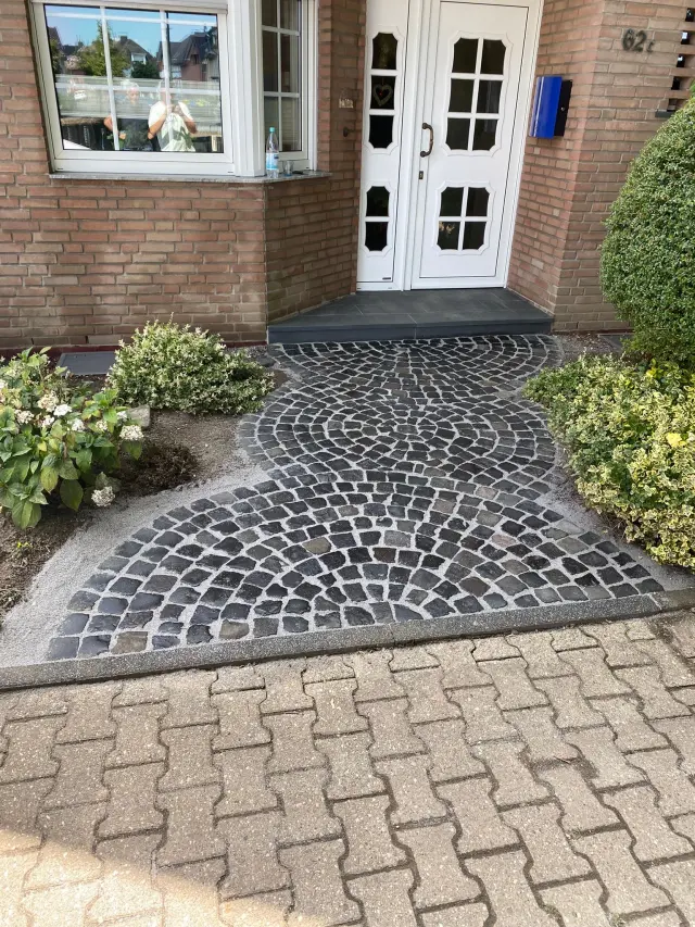 Sanierter Hauseingang mit Natursteinpflaster, gepflegten Beeten und weißer Haustür als Beispiel für Baulösungen und Sanierungslösungen.